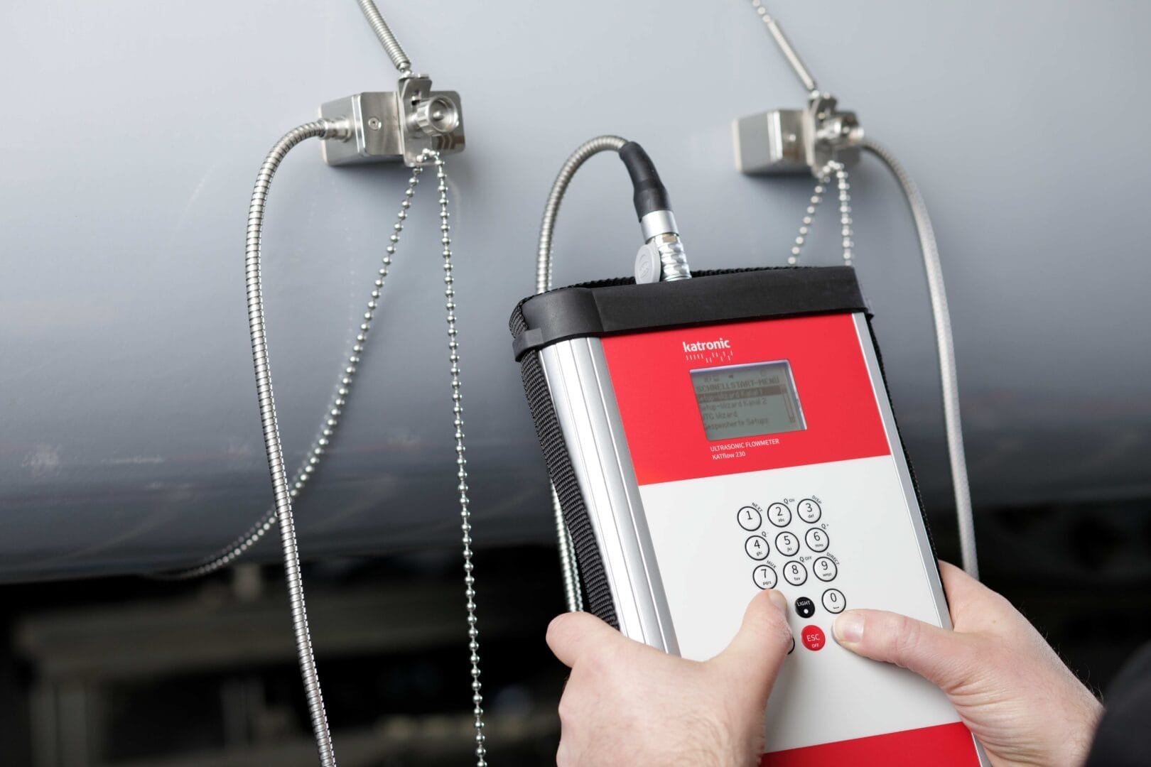 Person bruker et Katronic ultralyd‑flowmeter med clamp‑on sensorer montert på et rør.