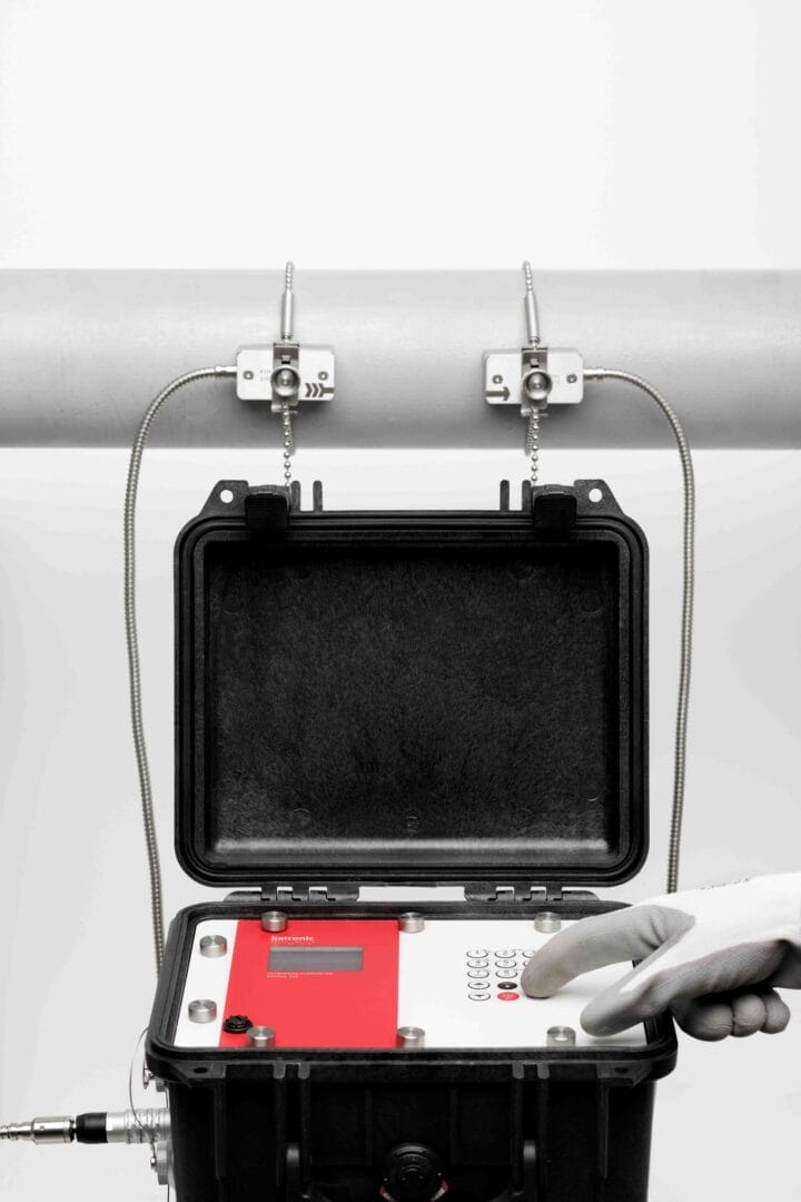 Katronic‑flowmeter i åpen transportkoffert koblet til to clamp‑on sensorer montert på et rør, med en hånd i hanske som betjener instrumentpanelet.