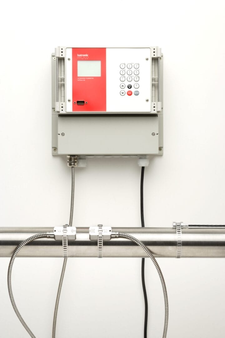 Fastmontert Katronic‑flowmeter med display og tastatur koblet til to clamp‑on sensorer montert på et metallrør.