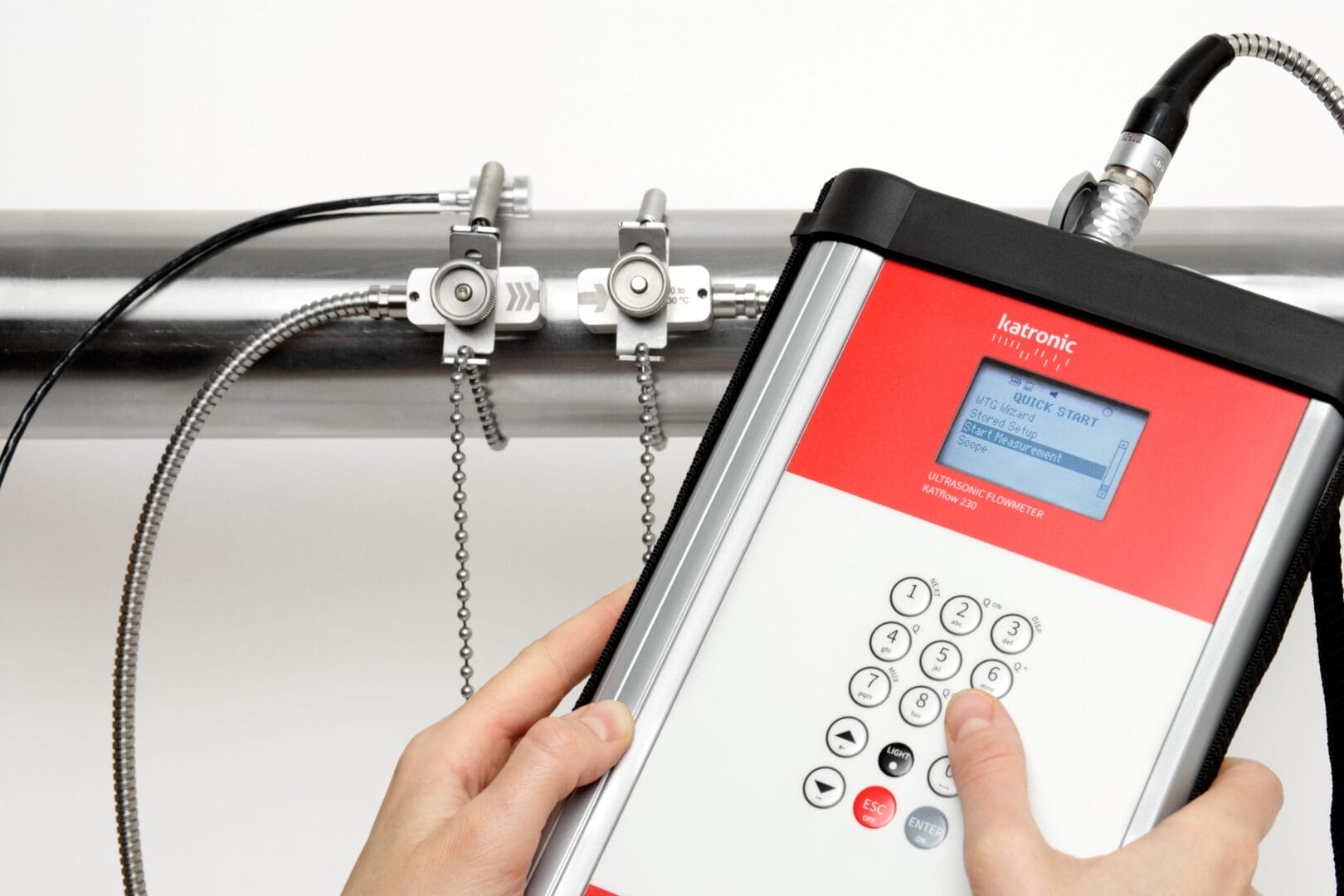 Person bruker et Katronic ultralyd‑flowmeter med clamp‑on sensorer montert på et rør.