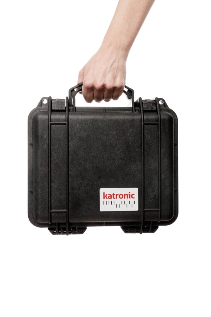 Hånd bærer en robust svart Katronic‑transportkoffert med logo på forsiden.