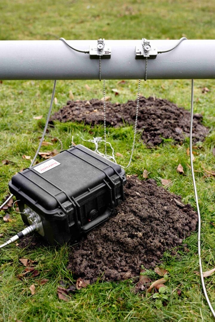 Portabelt Katronic‑flowmeter i en robust koffert koblet til to clamp‑on sensorer montert på et utendørs rør med kabler som går ned i gress og jord.